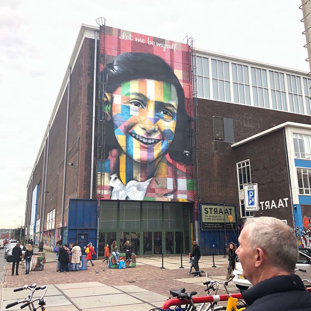 Colorful street art mural on a large building in Amsterdam, with people and bicycles in the foreground.