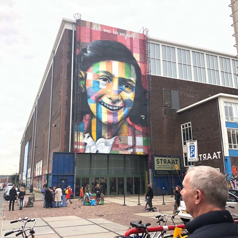 Colorful street art mural on a large building in Amsterdam, with people and bicycles in the foreground.