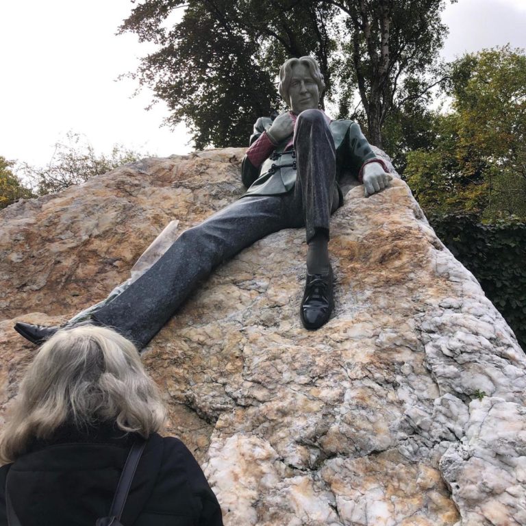 Statue of a seated figure on a rock with trees in the background, viewed by a gray-haired person.
