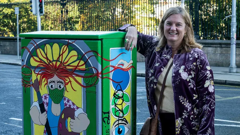 Woman smiling next to a vibrant street art display on a utility box in an urban setting.