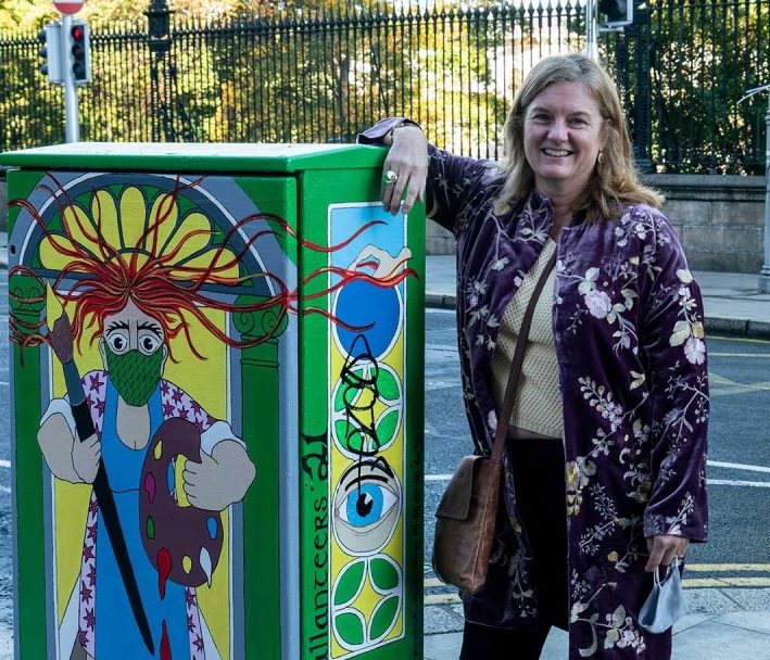 Woman smiling next to colorful painted utility box on city street.