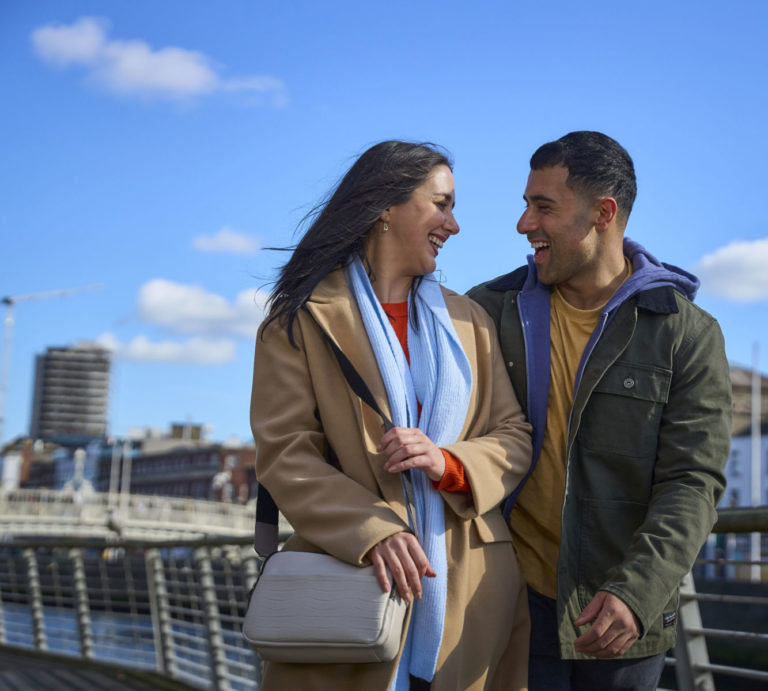 Couple laughing and walking on a bridge under a clear blue sky.