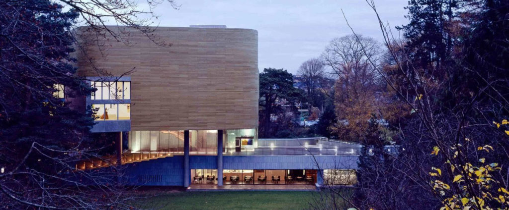 Modern building surrounded by trees at dusk, featuring unique architectural design and illuminated interiors.