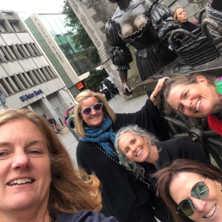 Group of women smiling near a bronze statue in a city street.