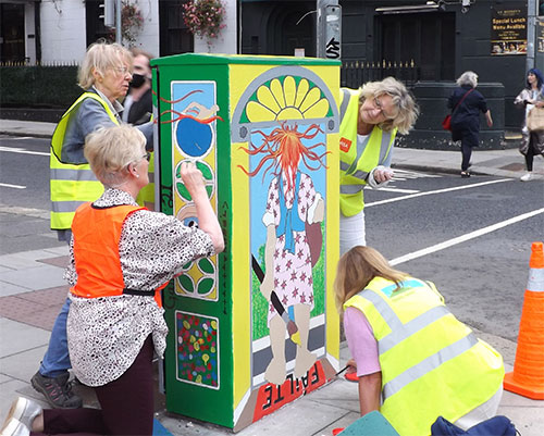 Artists in safety vests paint a colorful mural on a street utility box, enhancing urban landscape with vibrant art.
