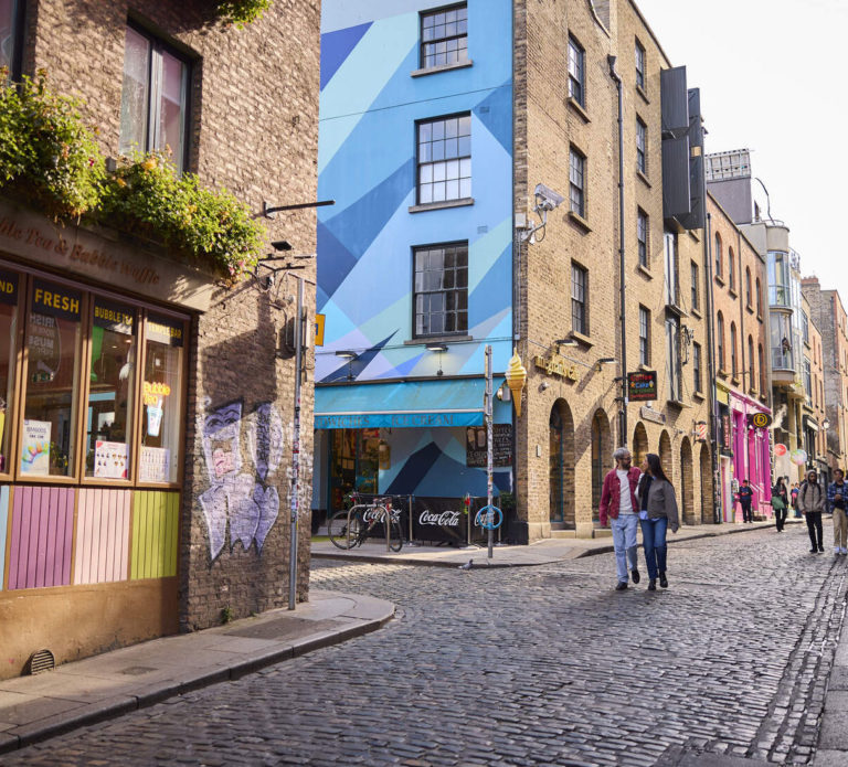 Colorful urban street with graffiti, vibrant shops, cobblestones, and people walking in a lively city setting.