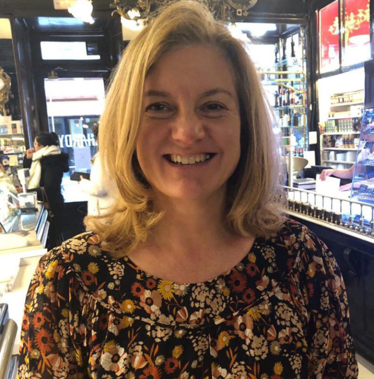 Smiling woman in floral top inside a store with display shelves and a person in the background.