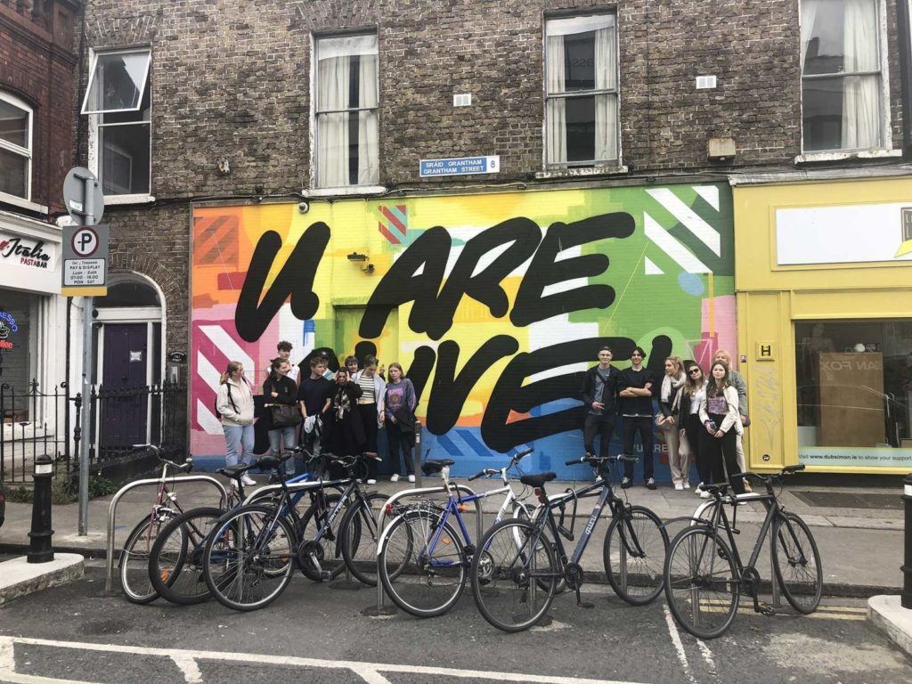 Street mural with vibrant colors and U ARE ALIVE text, people standing in front, bicycles parked nearby.