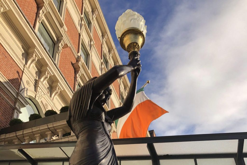 Statue holding torch with Irish flag background, beside ornate brick building under blue sky.