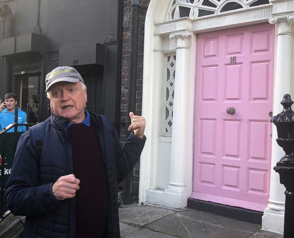 Man speaking outside a building with a bright pink door and decorative trim.