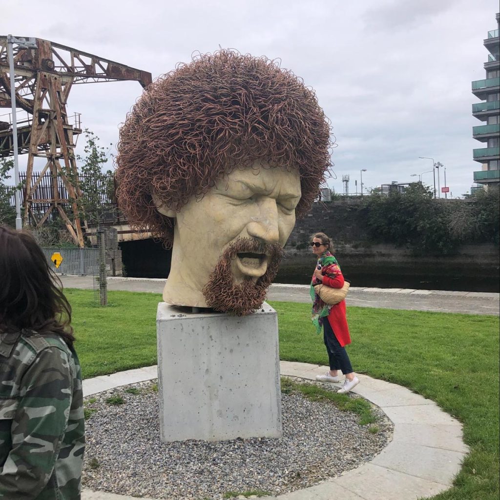 Sculpture of a large head with curly hair in a park setting, people nearby, industrial backdrop.