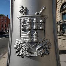 Silver emblem on a pole in a city street, featuring a detailed crest with architectural elements and a scroll design.
