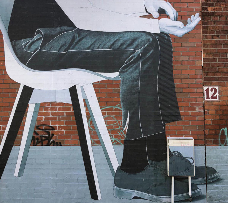 Mural on brick wall depicting a seated figure in a suit, number 12 visible on the wall, urban art scene.