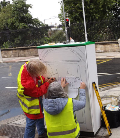 People painting a utility box on a street corner, wearing high-visibility vests, part of a street art project.