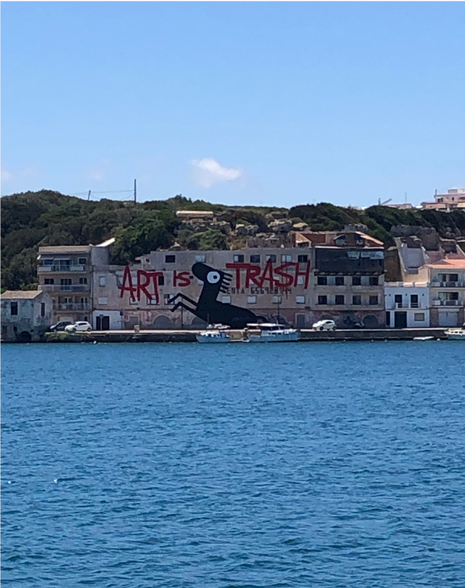 Street art mural on building reads Art is Trash featuring black figure, visible from water in scenic coastal setting.