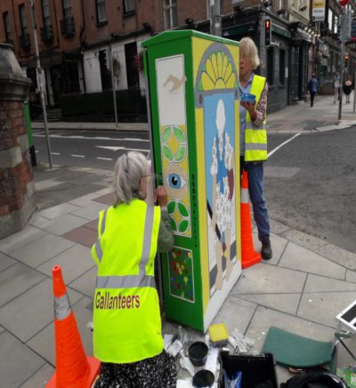 Artists in high-visibility vests painting mural on city electrical box, surrounded by traffic cones.