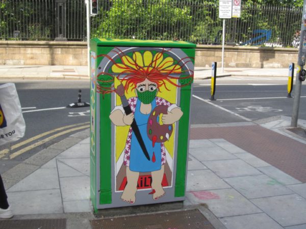 Colorful street art on utility box depicting a masked painter on an urban sidewalk.