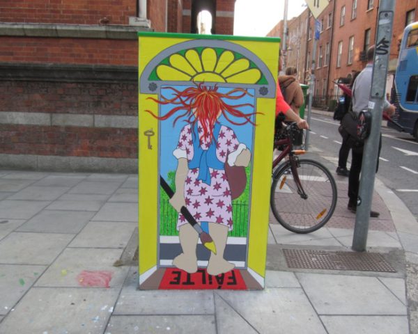 Street art on utility box shows a colorful figure under an arch. Urban scene with cyclist passing by.