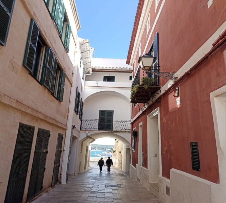 Narrow European street with colorful buildings, archway, and ocean view in the distance. Two people walk ahead.