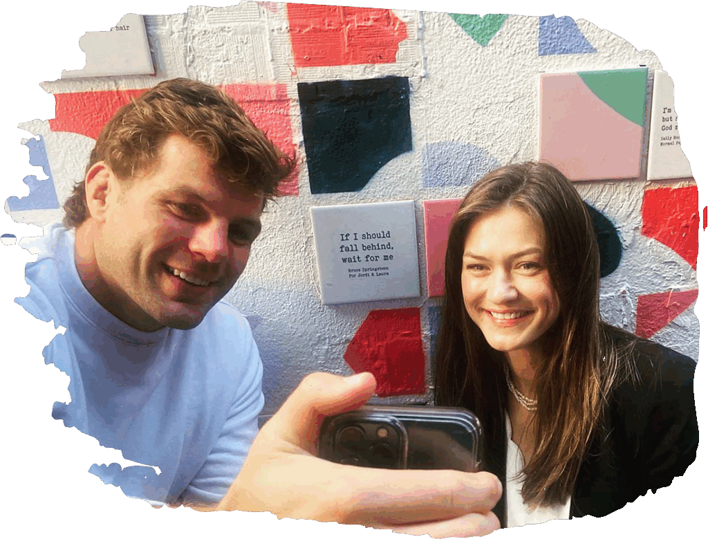 A couple taking a selfie against a colorful mural with inspirational quotes on the wall in the background.