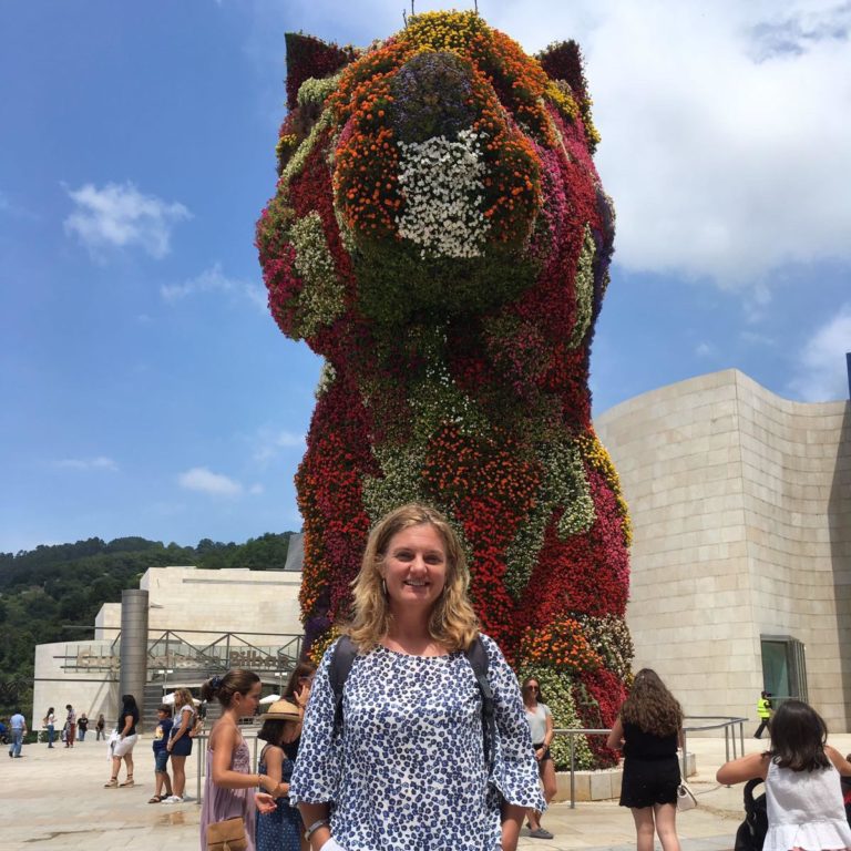 Smiling woman in front of colorful flower sculpture at Guggenheim Museum Bilbao, under a bright blue sky.