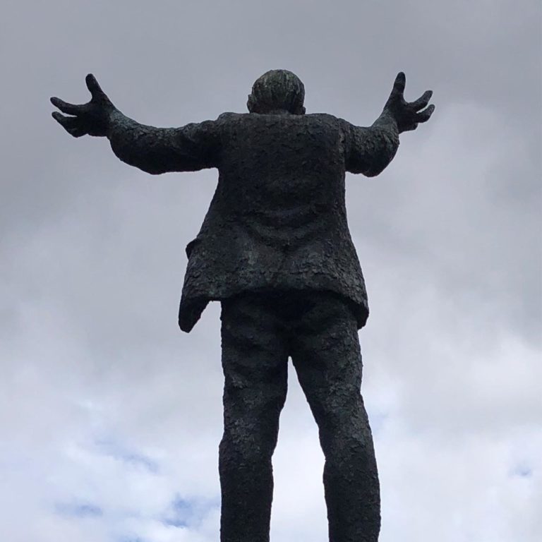 Bronze statue of a man with open arms, viewed from behind against a cloudy sky backdrop.
