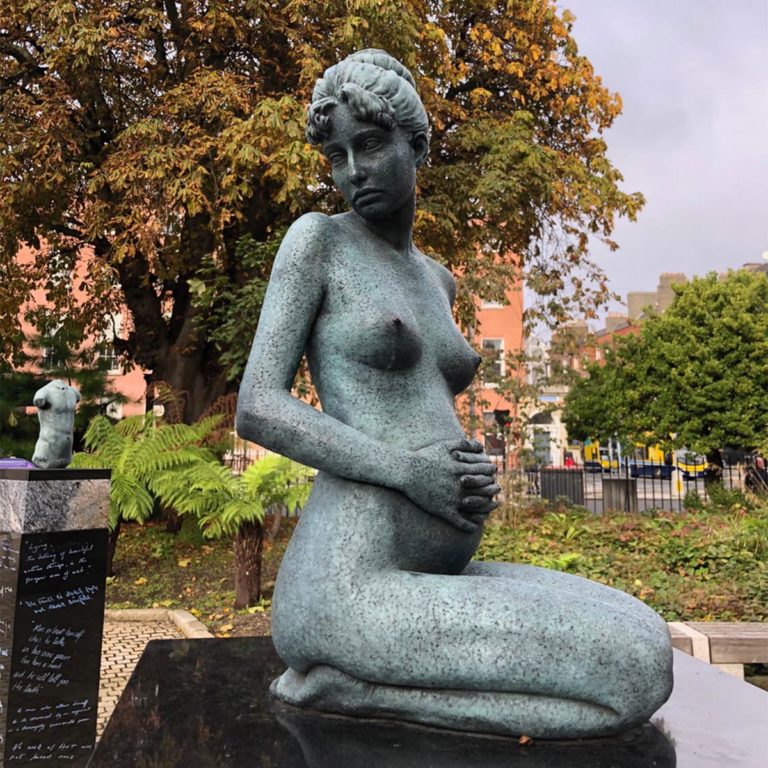Stone sculpture of a kneeling, pregnant figure in a park setting, surrounded by trees and buildings.