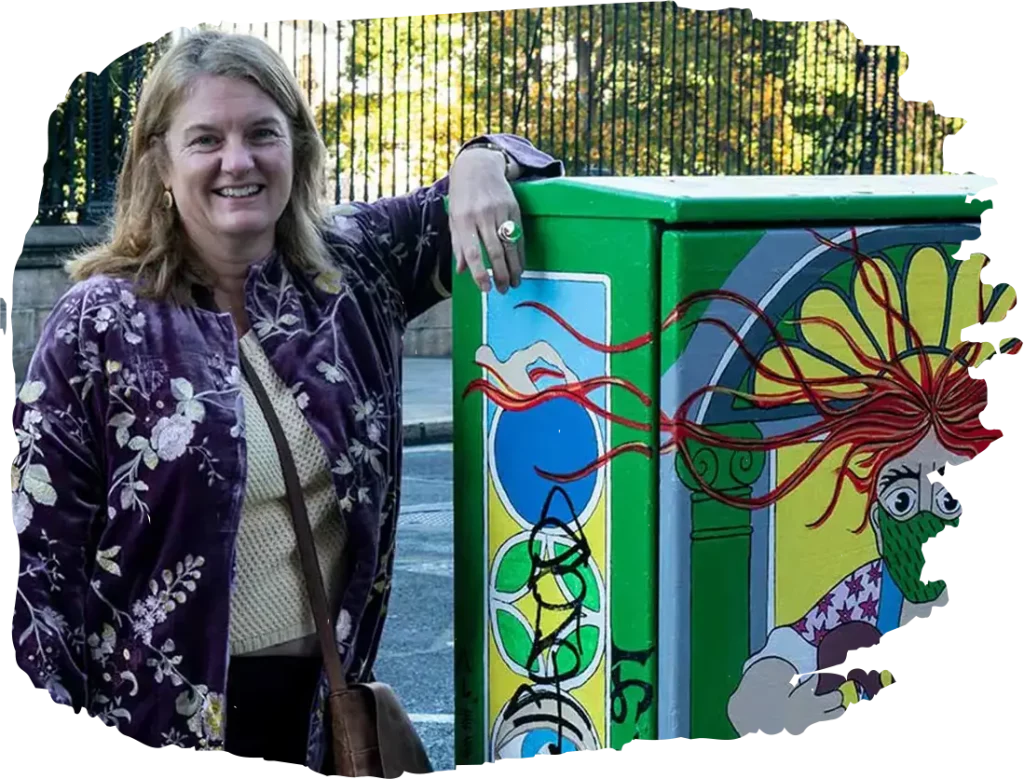 Smiling woman in floral jacket stands by colorful street art on utility box with abstract design.