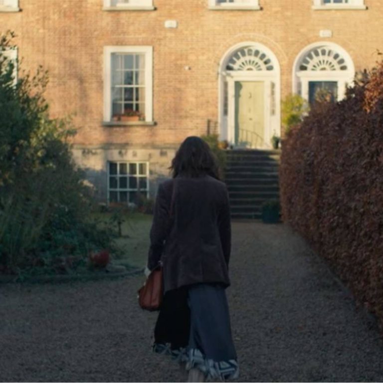 Person walking toward a brick house on a gravel path, carrying a bag, surrounded by hedges and greenery.