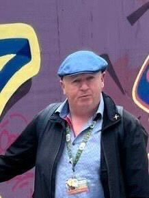 Man in a blue cap stands against a colorful graffiti wall, wearing a lanyard and black jacket.